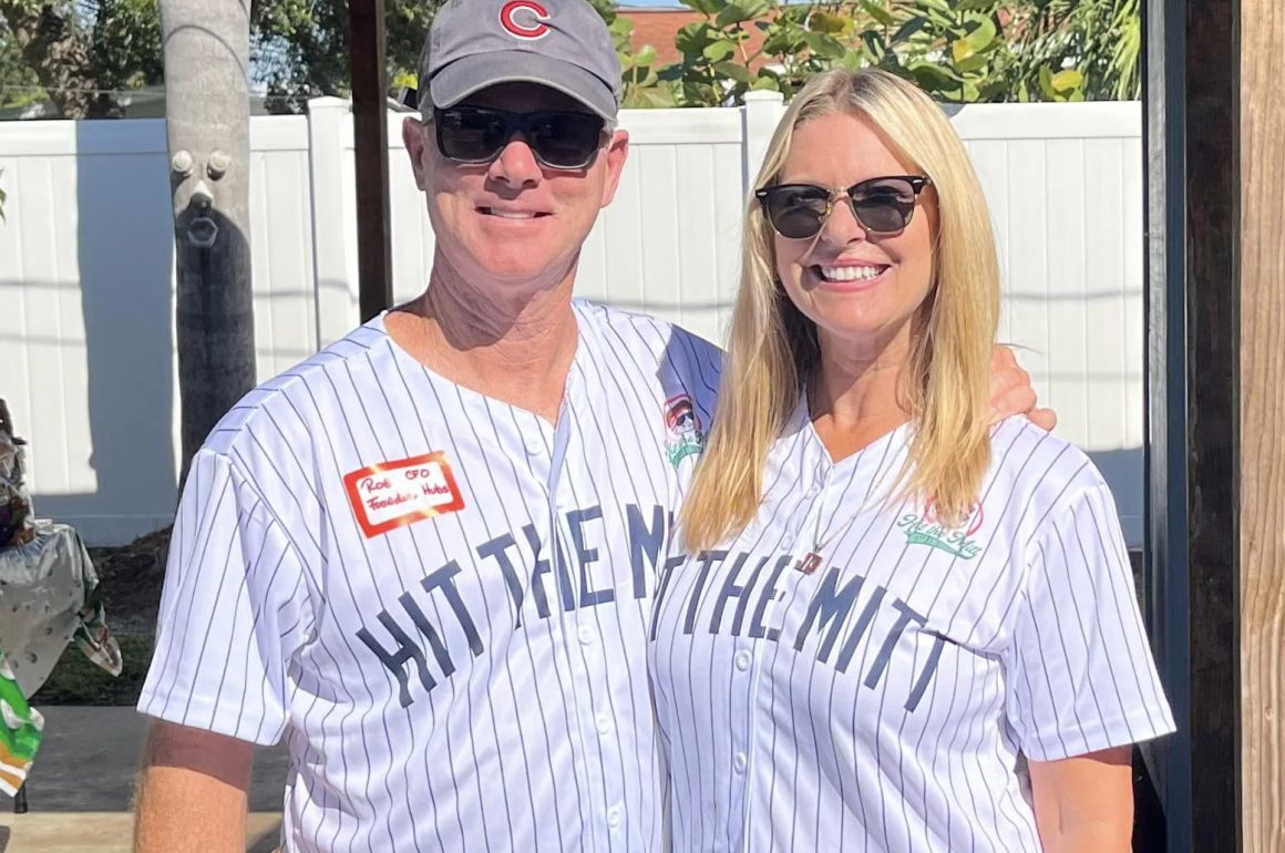 A man and a woman in baseball jerseys and sunglasses smile at the camera outside.