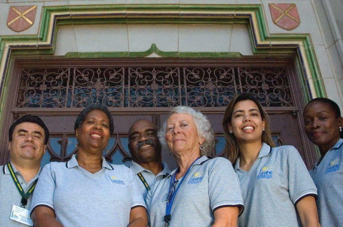 A photo looking up to a group of people in gray t-shirts smiling at the camera in front of a building.