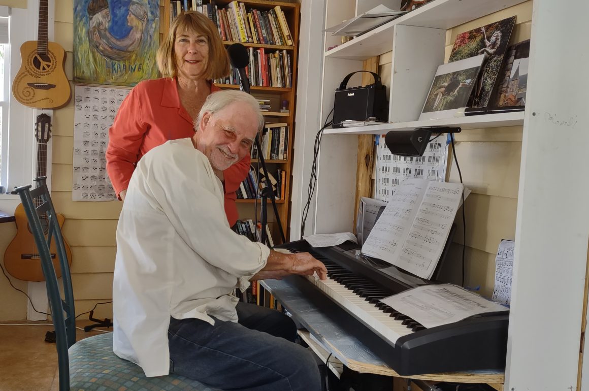 A photo of a woman standing behind a man playing piano while looking at the camera.